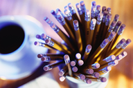 Sobre una mesa de madera, visto desde arriba, junto a una taza de café solo, otra taza contiene gran cantidad de lápices iguales.